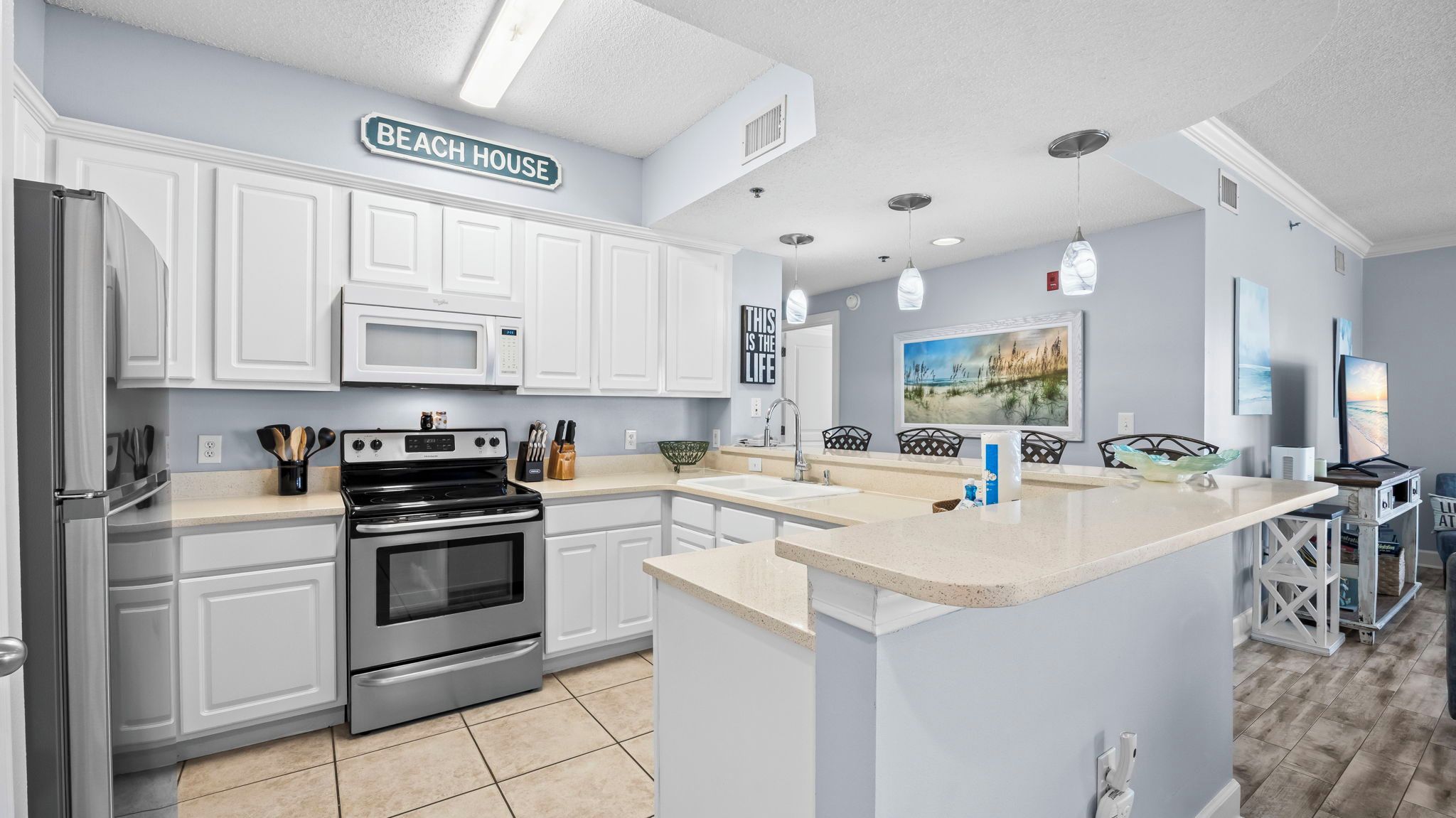 A kitchen with white cabinetry, a stainless steel refrigerator, a microwave, and an oven. There is a sign above the cabinets that reads 'BEACH HOUSE.'