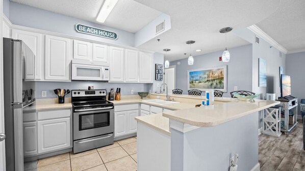 A kitchen with white cabinetry, a stainless steel refrigerator, a microwave, and an oven. There is a sign above the cabinets that reads 'BEACH HOUSE.' - Palm Beach B31 Gulf Front 3BR 2BA Corner Unit (Orange Beach)