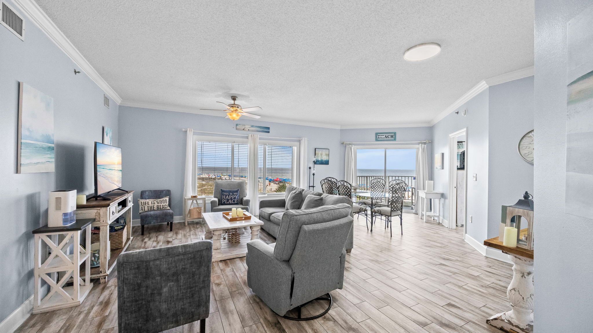 A spacious living room with a large window overlooking the beach, a ceiling fan, and a dining area with outdoor seating.