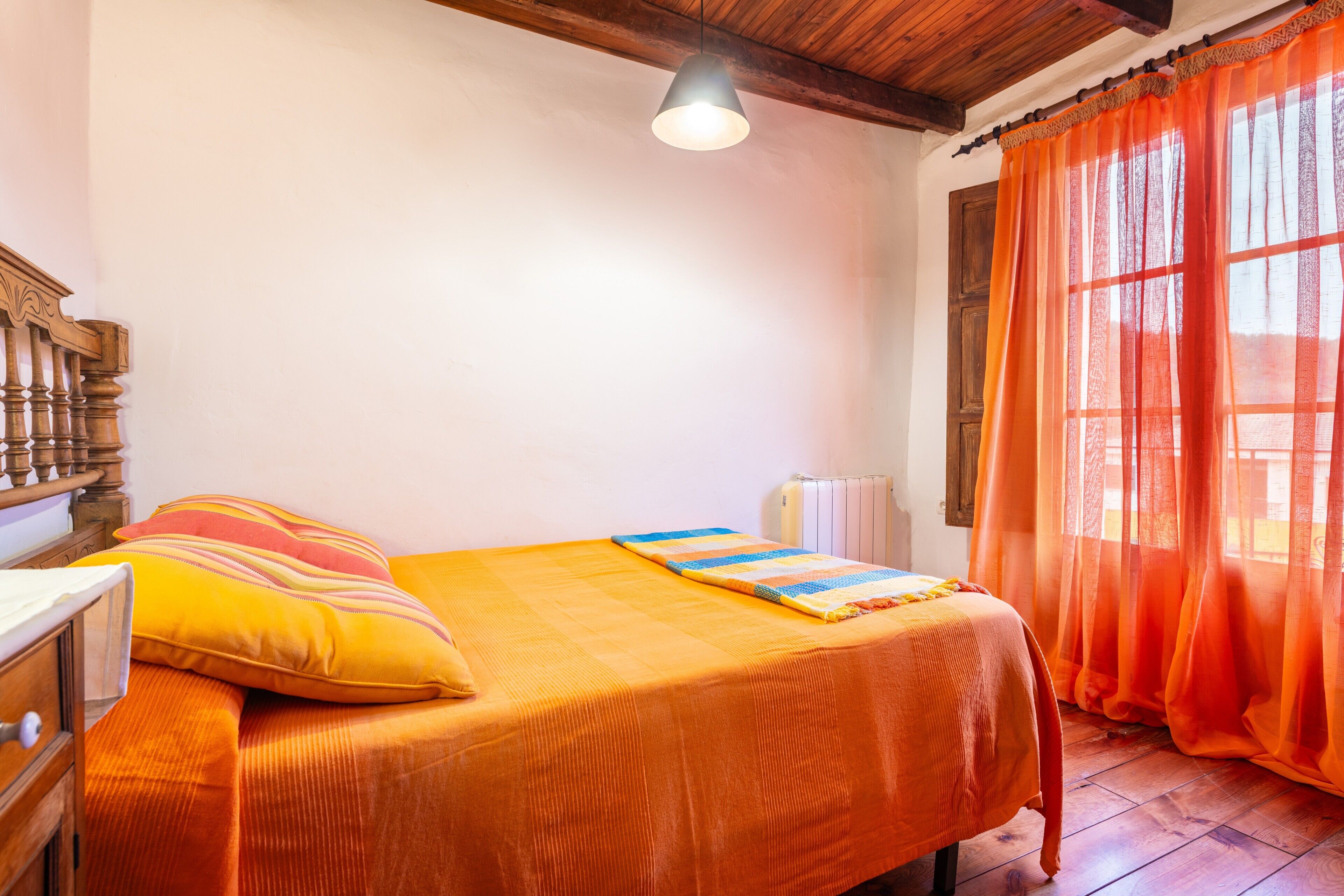 Un dormitorio con una cama, cabecero de madera, una ventana con cortinas naranjas y un radiador.