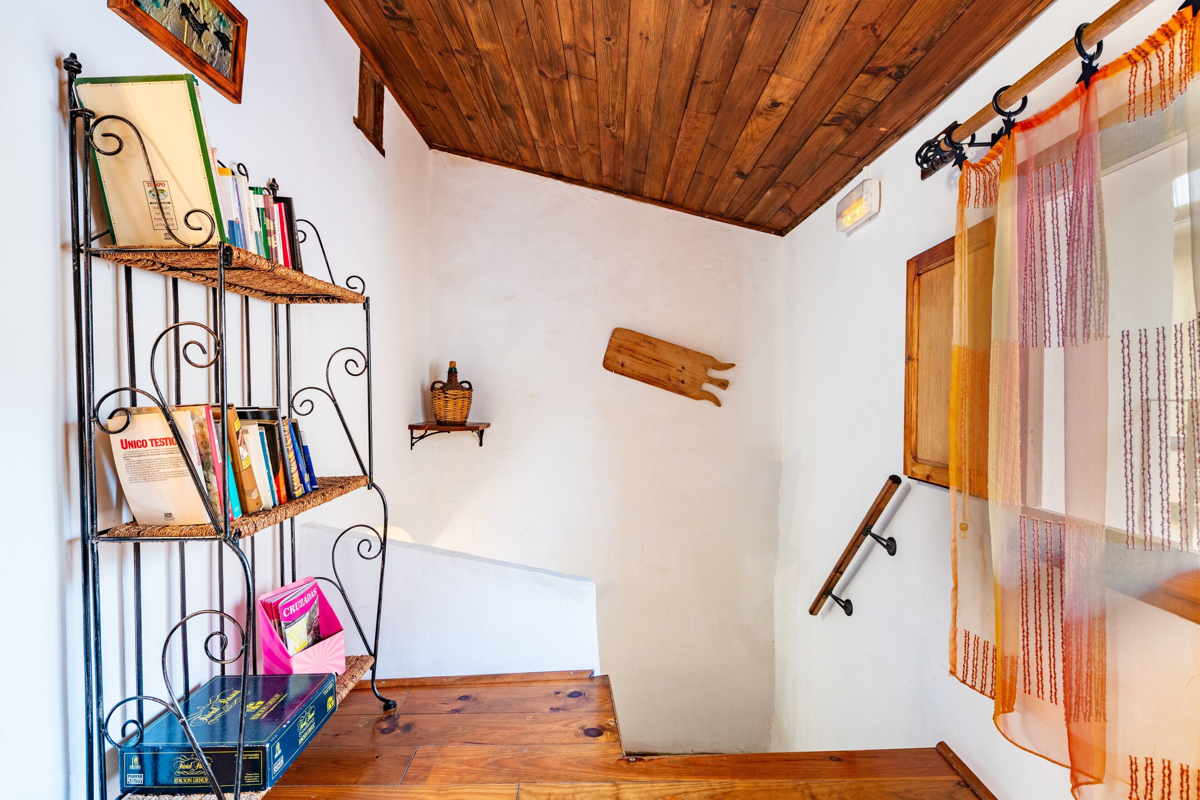 A corner of a room with a wooden ceiling, a metal bookshelf with books, a wicker basket, and a wooden cutting board on the wall.