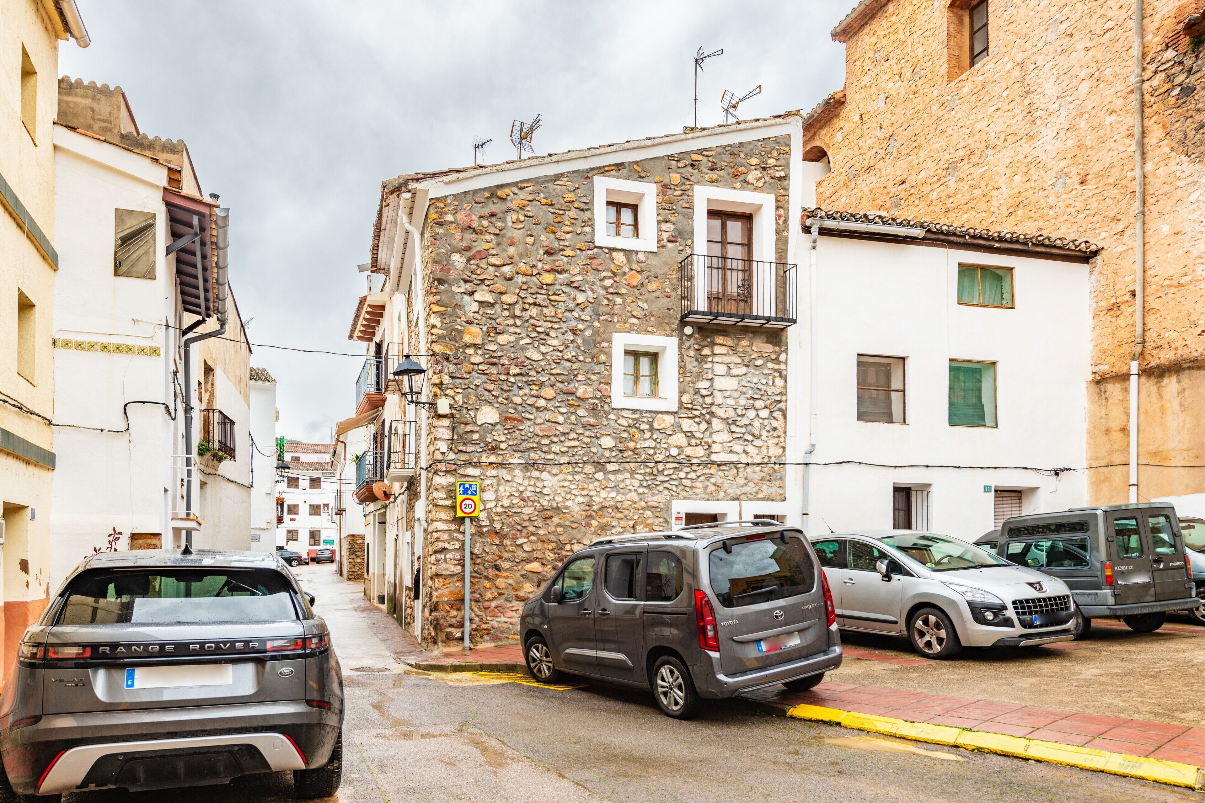 Una calle estrecha flanqueada por edificios de piedra, coches aparcados y una señal que indica un límite de velocidad de 20 km/h.