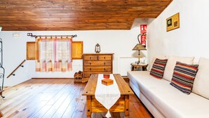 Une pièce avec un plafond en bois, une table en bois recouverte d’une nappe blanche, un canapé agrémenté de coussins à rayures, une commode en bois et une fenêtre avec des rideaux.