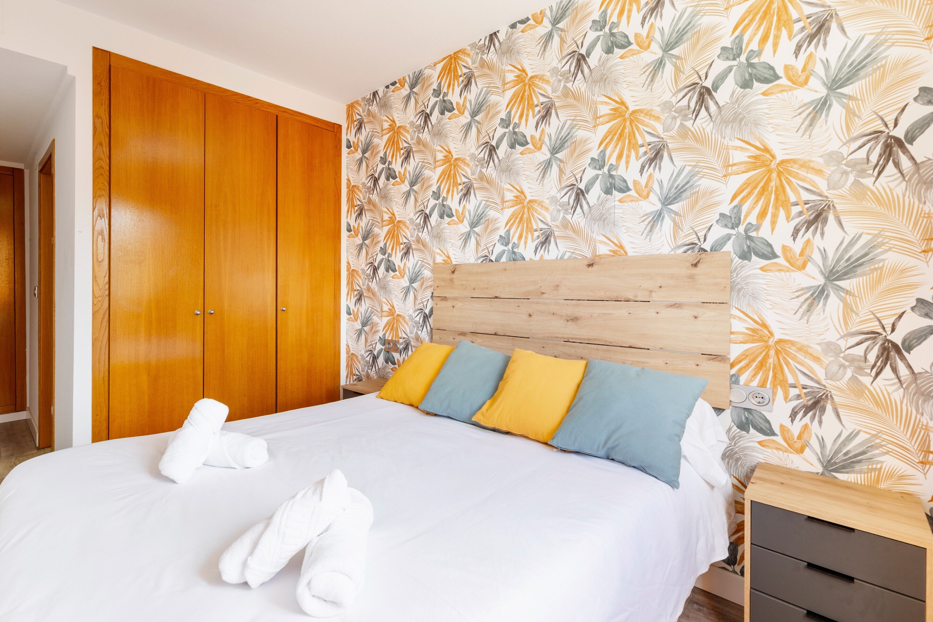 Habitación de hotel con un armario de madera, cama con ropa de cama blanca y pared con estampado de hojas tropicales.