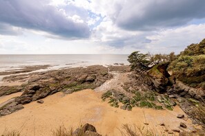 Ein felsiger Strandabschnitt mit Sandfläche und Blick auf das Meer unter einem bewölkten Himmel.