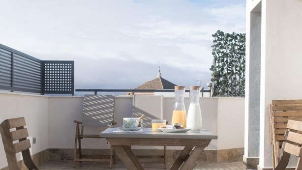 Un patio con una mesa lista para el desayuno, dos sillas de madera, una parrilla metálica y vista al mar.