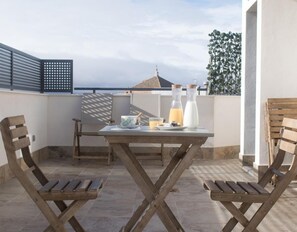 Un patio con una mesa lista para el desayuno, dos sillas de madera, una parrilla metálica y vista al mar.