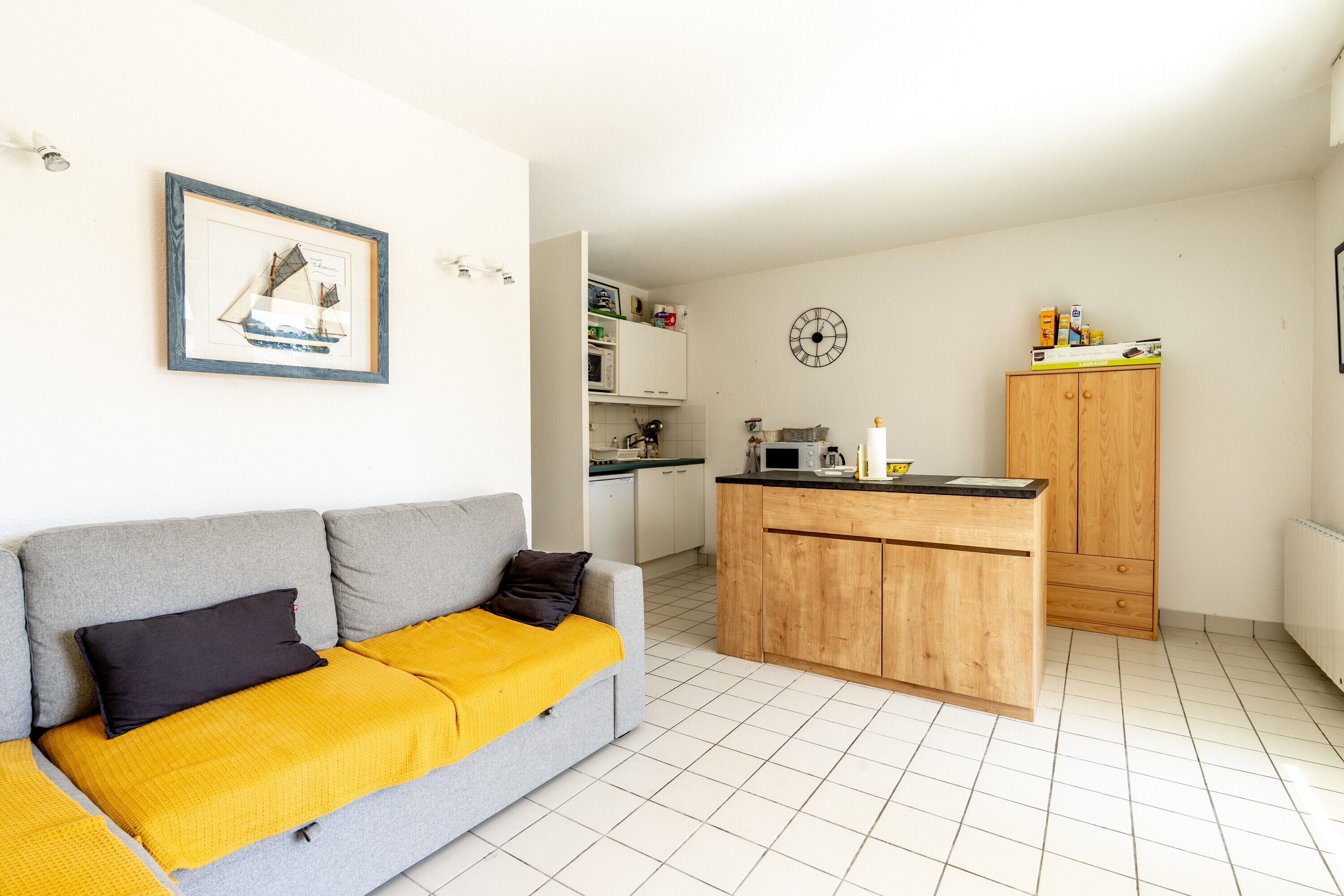 Un espacio habitable compacto con kitchenette, un sofá con una manta amarilla y un cuadro en la pared.