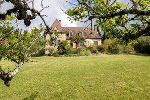 Une maison en pierre avec une grande pelouse, entourée d’arbres et d’arbustes.
