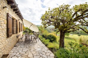 Une terrasse en pierre avec des meubles d’extérieur, un grand parasol et un arbre.