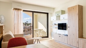 Un salon moderne avec un canapé, un fauteuil rouge, une table à manger, un téléviseur et une porte-fenêtre coulissante donnant sur un balcon.
