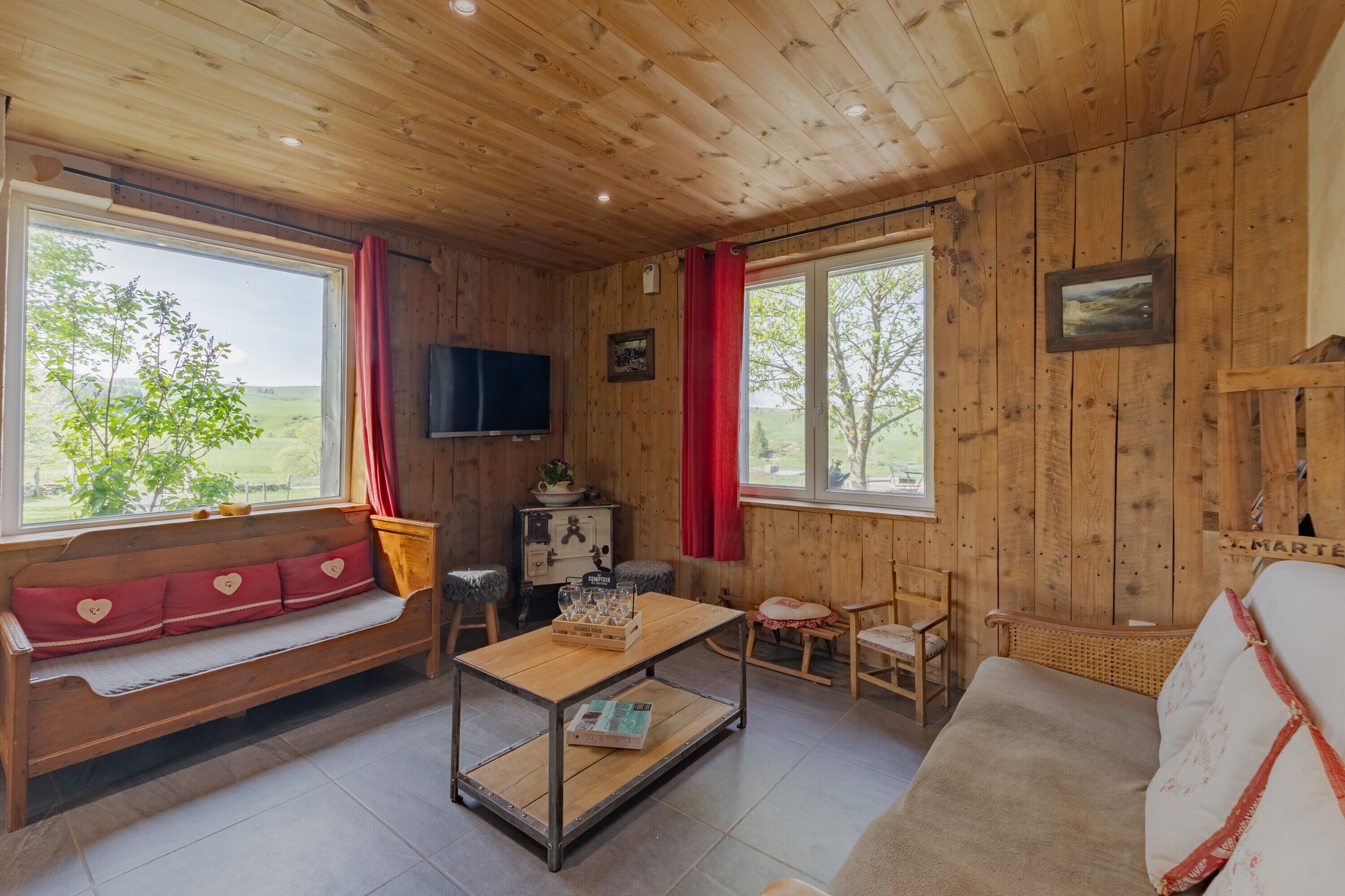 Een houten hut van binnen, met een houten bank, een tafel met glazen, een televisie en uitzicht op de natuur.
