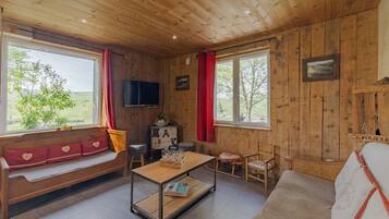 Een houten hut van binnen, met een houten bank, een tafel met glazen, een televisie en uitzicht op de natuur.