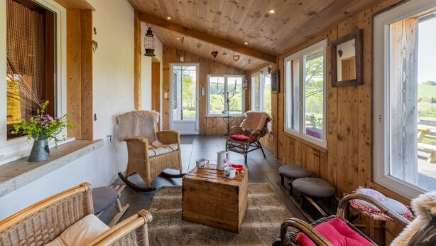 A cozy room with wooden walls and ceiling, wicker furniture, and large windows offering a view of the outdoors. - Discover Gîte Le Pradel in Les Estables
 (Les Estables)