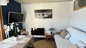 A modern living room with a flat-screen TV, a sofa, a dining table with chairs, and a large window.