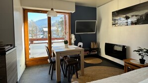 A modern bedroom with a bed, a TV, a dining area with a table and chairs, and a view of mountains through the window.
