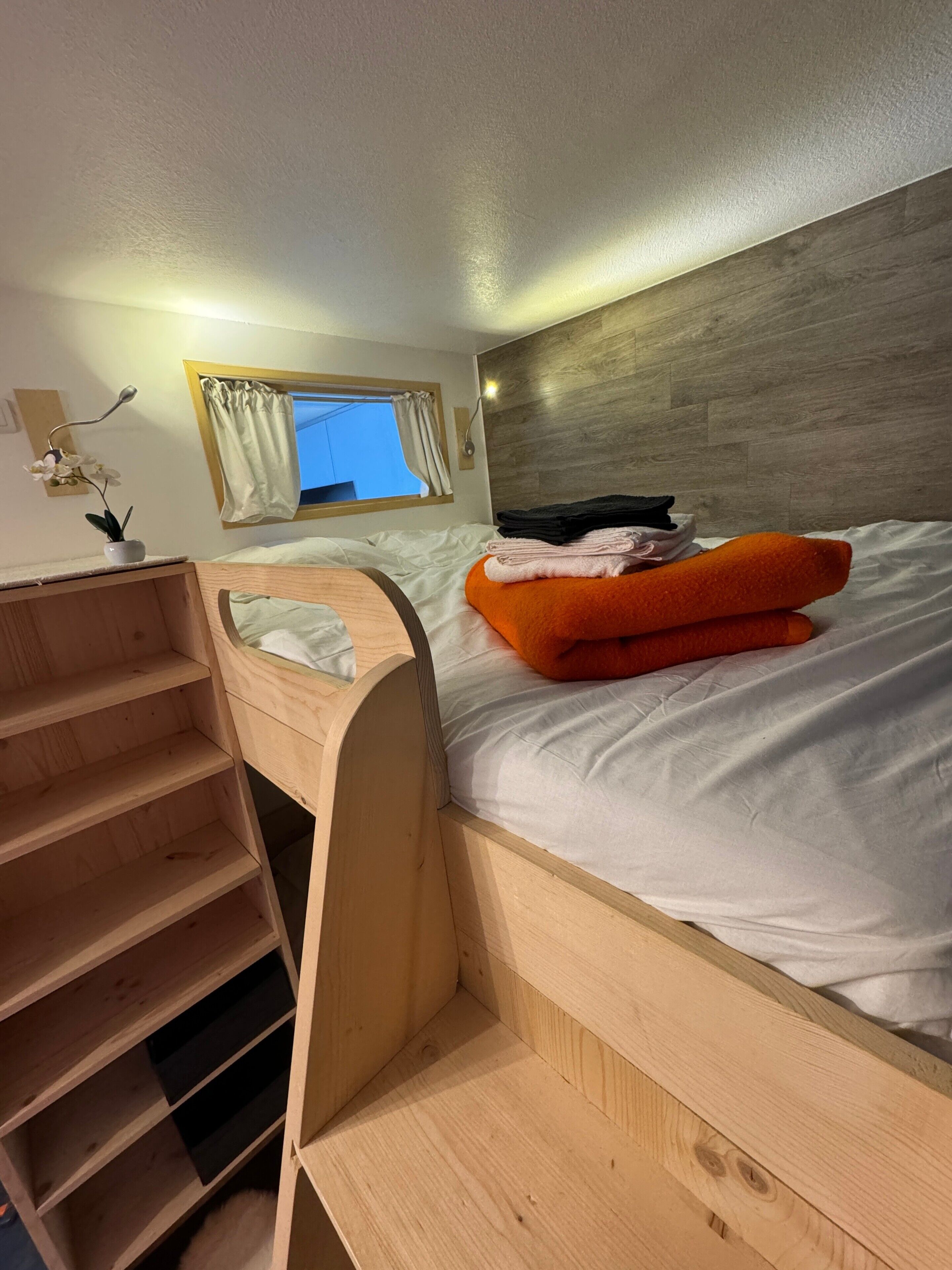 En køjeseng med trægavl, et natbord med lampe og et vindue med gardiner.