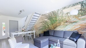 Een moderne woonkamer met een grijze hoekbank, een fauteuil met een patroon, een eethoek met een witte tafel en stoelen, en een trap.