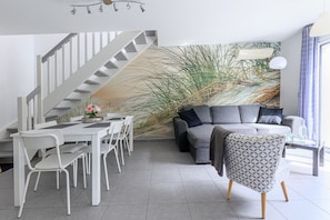 A modern living room with a staircase, grey sofa, white dining table, and chairs.
