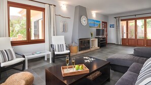 A living room with a sofa, chairs, a coffee table with a tray of apples and glasses, a fireplace, and a view through large windows. - Country House 'Ses Penyes' with Sea View, Wi-Fi and Air Conditioning (Santanyí)