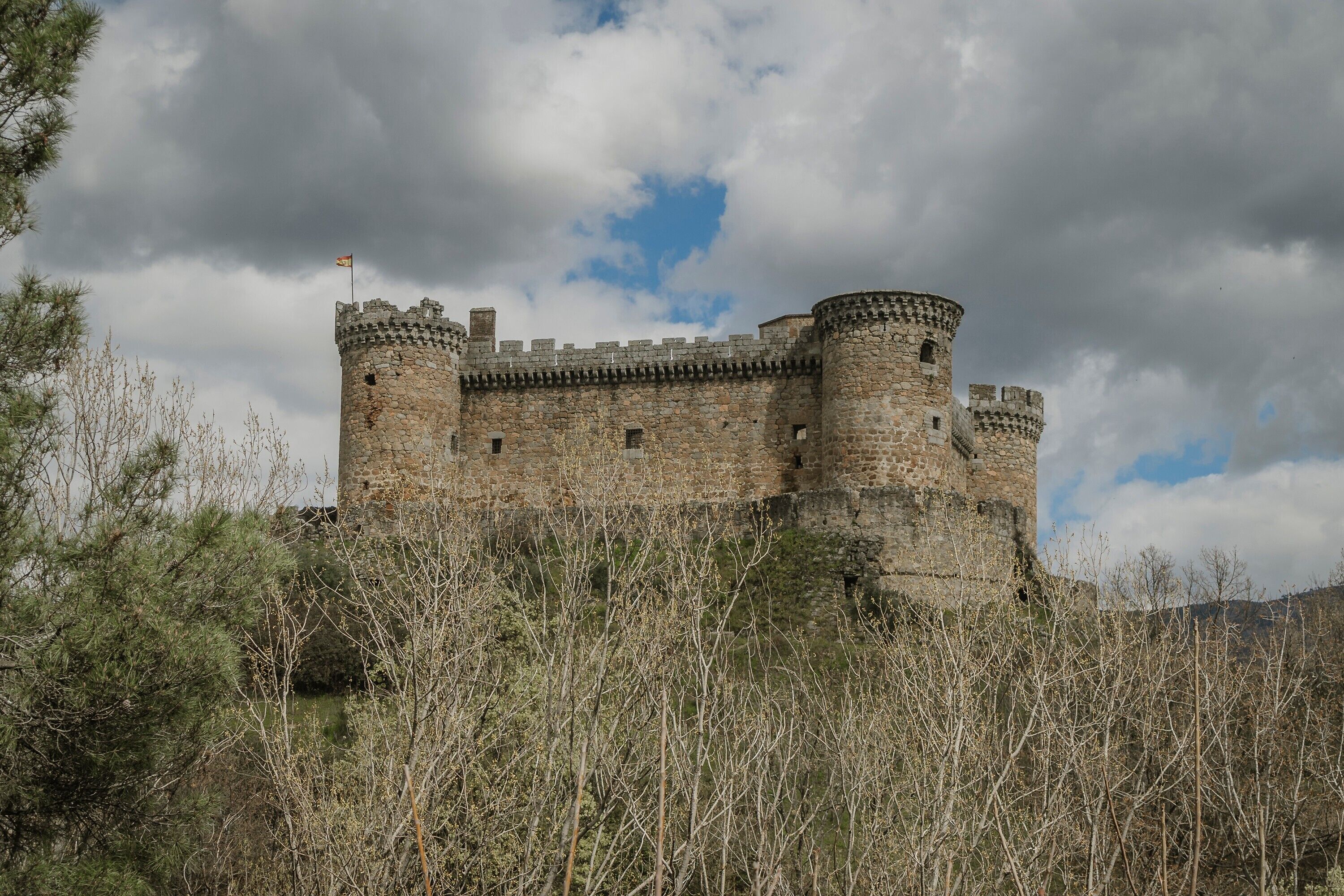 Un château de pierre historique, doté de nombreuses tours et surmonté d’un drapeau, entouré d’une végétation dense.