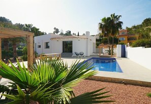 A modern villa with a swimming pool, a patio with seating, and a garden with palm trees.