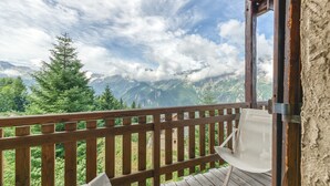 Une terrasse en bois avec une chaise blanche, offrant une vue sur un paysage montagneux parsemé d’arbres et de nuages.