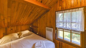 Une chambre dans une cabane en bois, avec un lit, une fenêtre ornée de rideaux en dentelle et un radiateur.