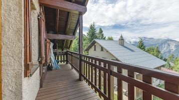 Eine Holzterrasse mit Geländer, ein Gebäude mit geneigtem Dach und eine gebirgige Landschaft im Hintergrund.