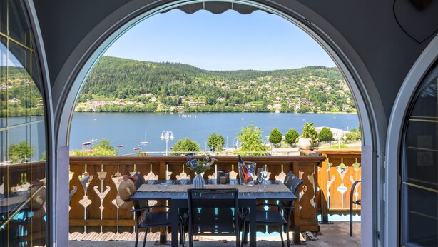 Une terrasse avec une table et des chaises, une vue sur un lac et une colline boisée.