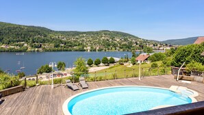 Une terrasse en bois avec une piscine, donnant sur un lac où l’on aperçoit des bateaux, et sur une ville en arrière-plan.