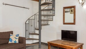 Una sala de estar con un sofá marrón, una mesa de centro de madera, un televisor, una escalera en espiral y un espejo fijado a la pared.