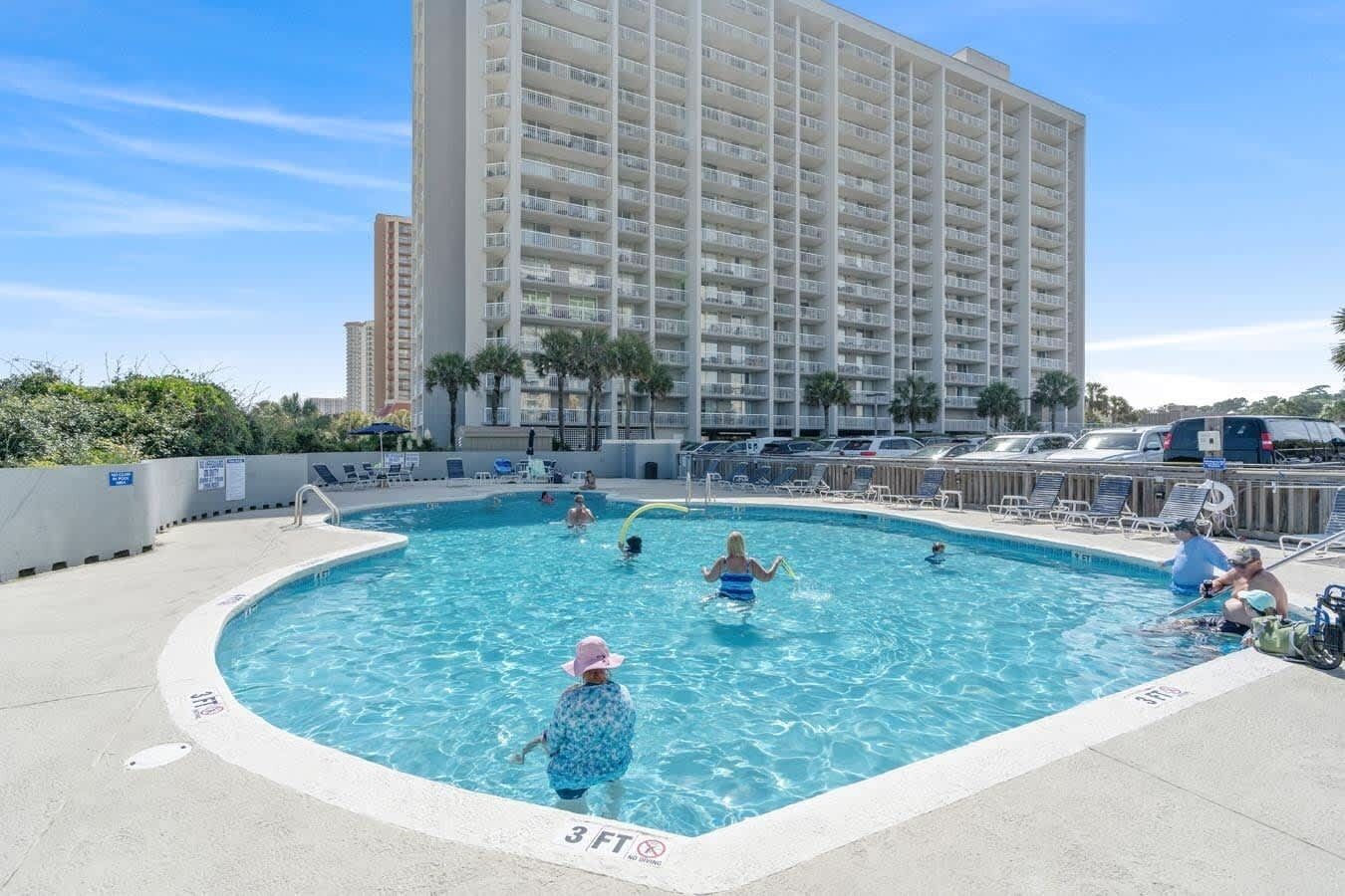 A swimming pool with people, a tall building in the background, and a clear sky.