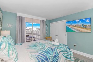 A bedroom with a large bed, a cityscape painting, a star decoration, and a view of a building complex through the window. - North Hampton 1607 (Myrtle Beach)