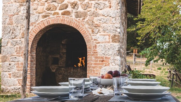 An outdoor dining area with a rustic brick oven, a wooden table set with plates, glasses, and a bowl of fruit.