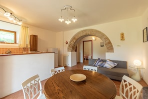 Un espace repas avec une table ronde en bois, des chaises blanches, un coin cuisine avec un comptoir, et un espace de vie avec un canapé et une lampe.