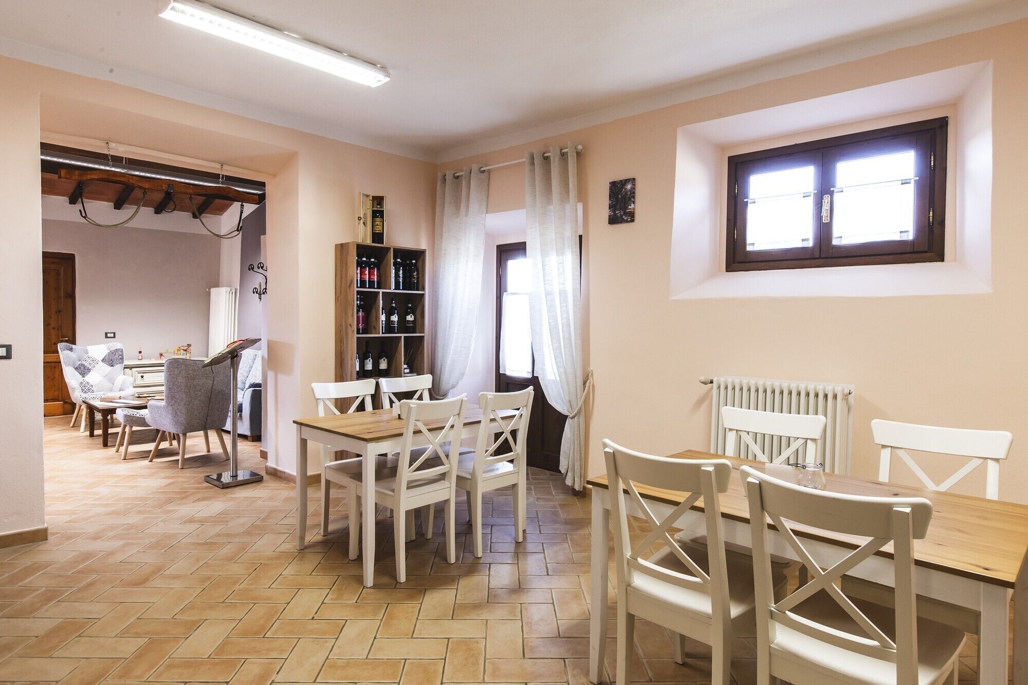 Un comedor con una mesa de madera y sillas blancas, una estantería con diversos objetos, y una ventana con cortinas.