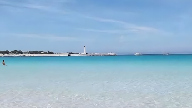 A beach with clear water, a lighthouse, and a boat in the distance.