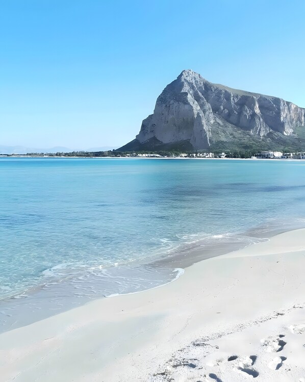 Uma praia com águas cristalinas, uma montanha imponente ao fundo e alguns edifícios ao longo da costa.
