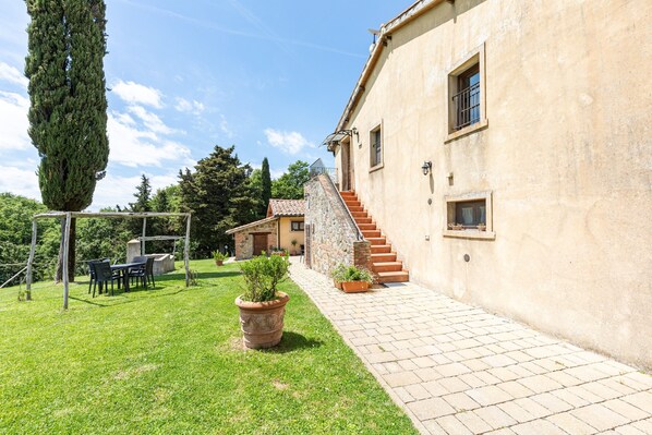 Un bâtiment en pierre avec un escalier menant à une porte, une zone herbeuse avec une table et des chaises, et un grand arbre.