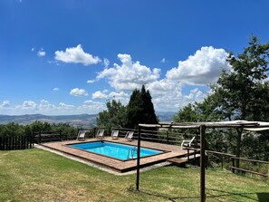 Piscina retangular com deck de madeira, cercada por uma vedação de madeira, com vista para uma paisagem pitoresca com árvores e colinas ao longe.