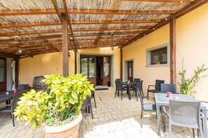 Um pátio externo com mesas e cadeiras, um vaso de planta e uma estrutura de pérgola de madeira.