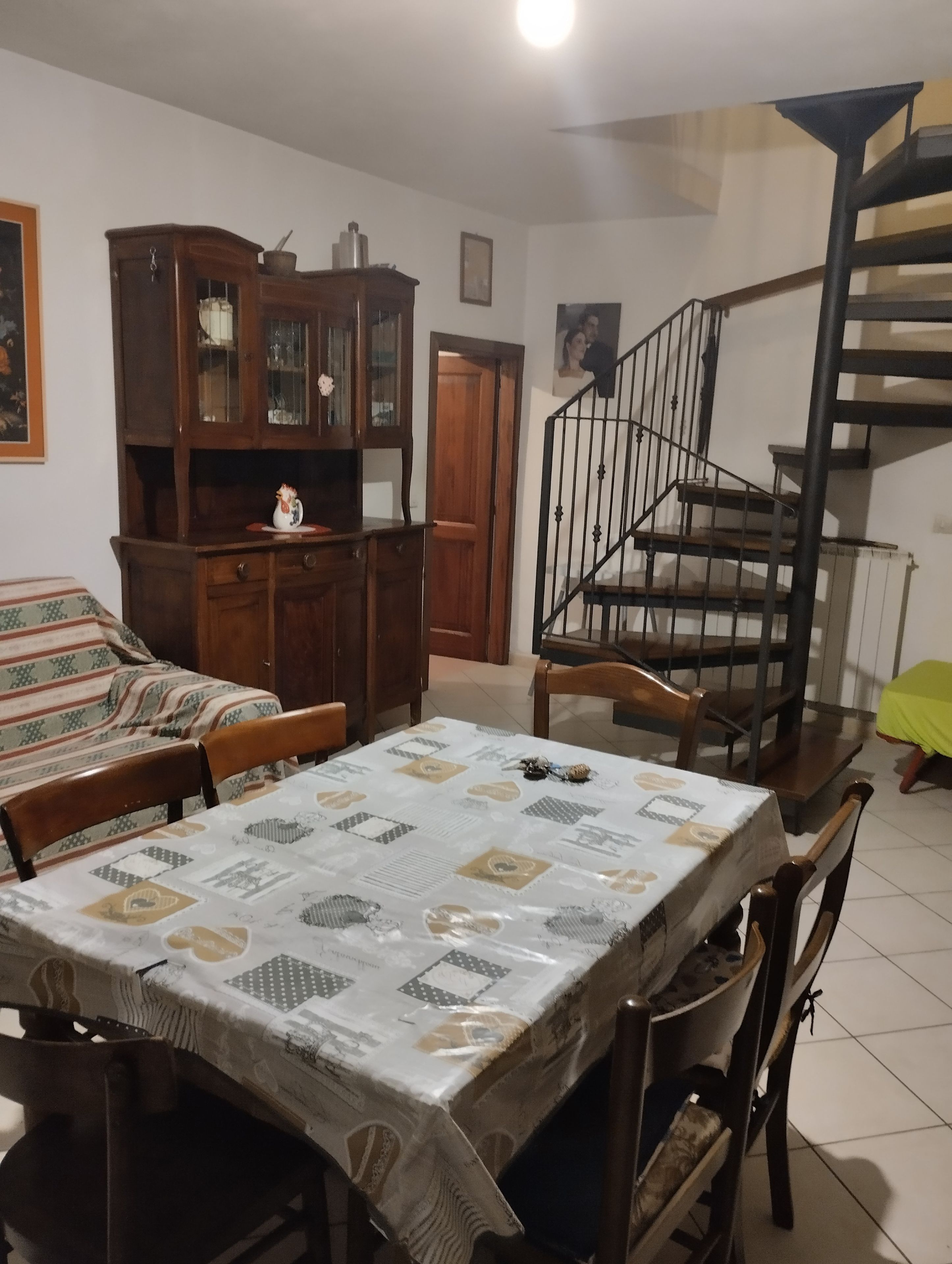 A dining area with a table set for four, a wooden cabinet with glass doors, a staircase, and a patterned tablecloth.
