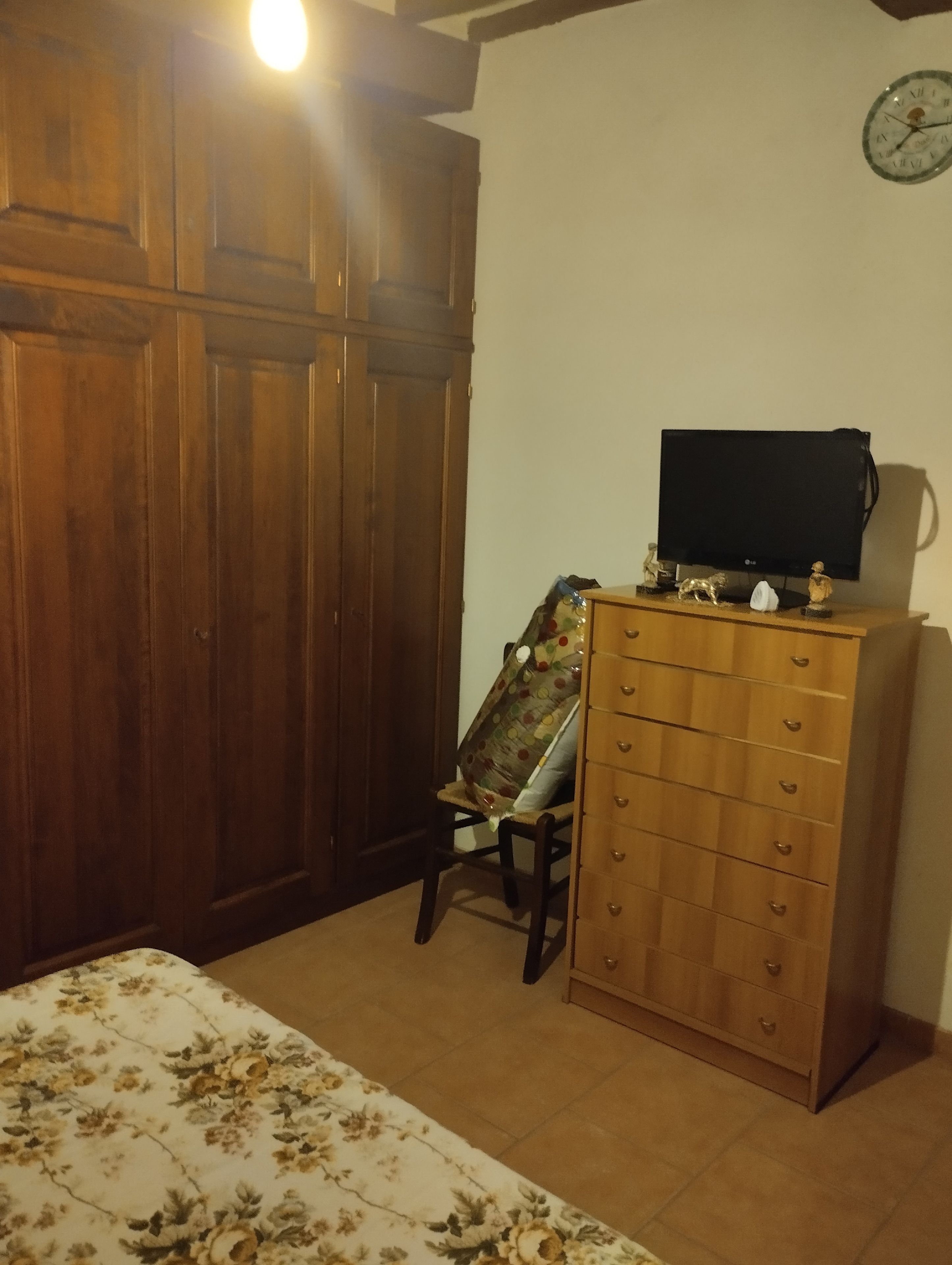 A room with a wooden wardrobe, a wooden dresser, a chair with a floral cushion, and a bed with a floral bedspread.
