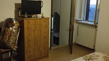Une chambre à coucher comprenant un lit, une commode, un téléviseur, une horloge, une fenêtre et une porte donnant sur une autre pièce.