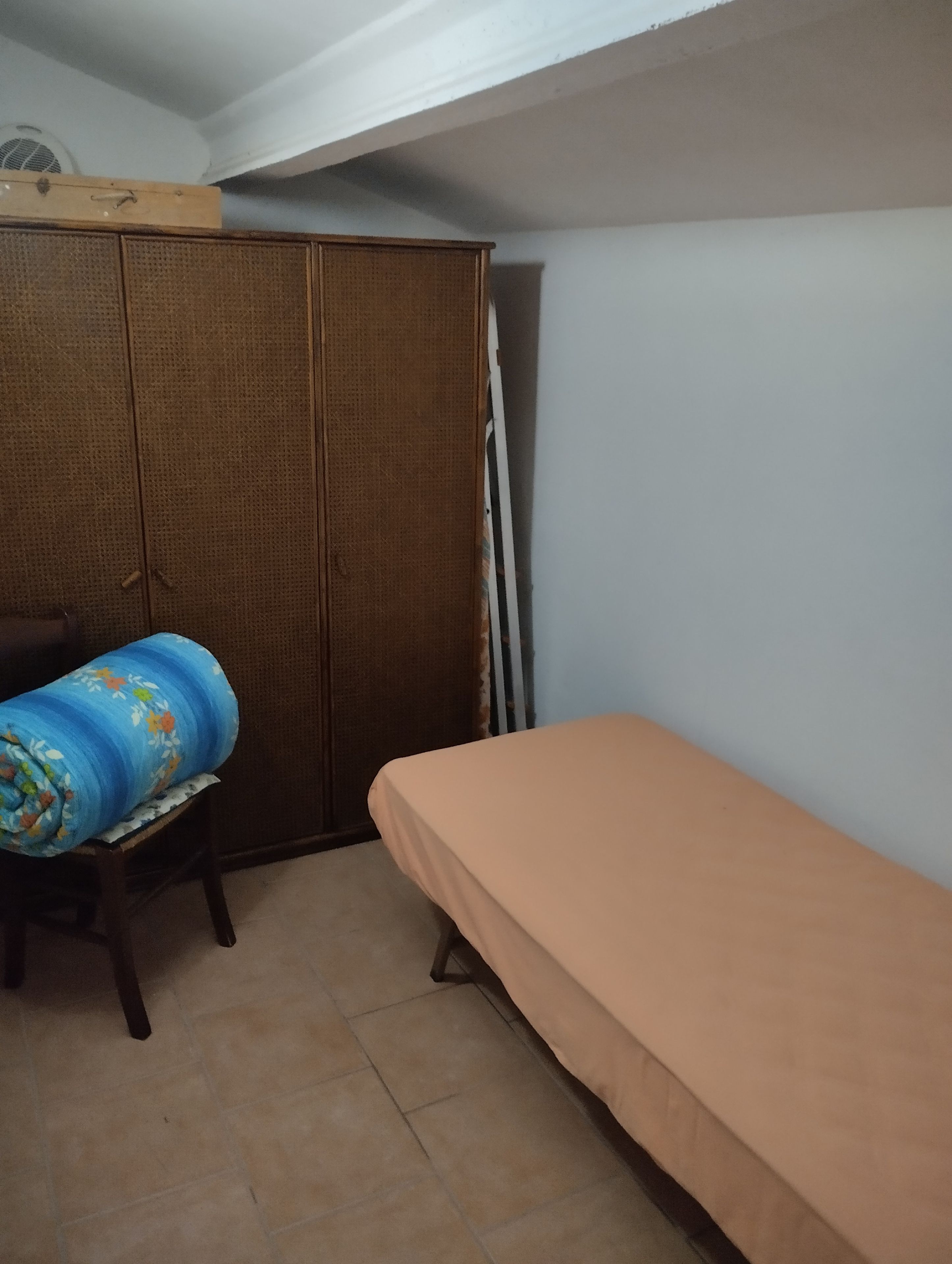 A small room with a bed, a wardrobe, and a chair with a rolled-up mat.