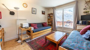 A cozy living room with a sofa, a coffee table, a bookshelf, a radiator, and a window with a view of mountains. - Apartment 'Les Arolles' with Mountain View, Shared Pool and Balcony (Valloire)