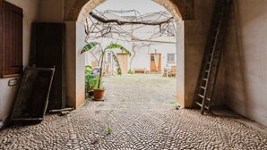 Un portail en arc donnant sur une cour dallée de pierres, avec une échelle et un pot de fleurs.