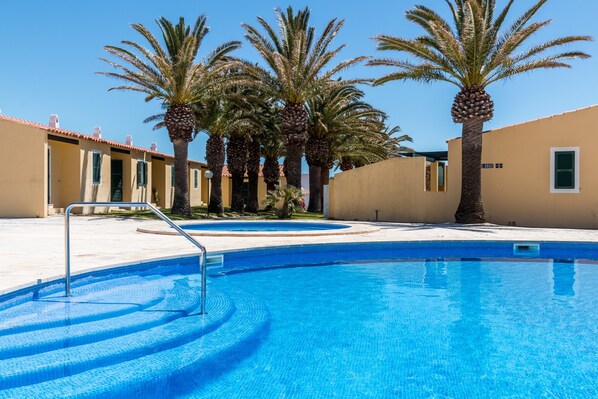 A swimming pool with clear blue water, surrounded by palm trees and beige buildings.