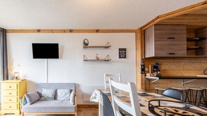 Un salon chaleureux avec un canapé, un téléviseur, des étagères en bois et un coin repas comprenant des chaises et une table.